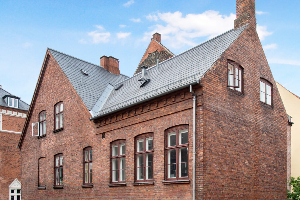 To ældre røde murstensbygninger med mørke tage og tagrender set mod en blå himmel.