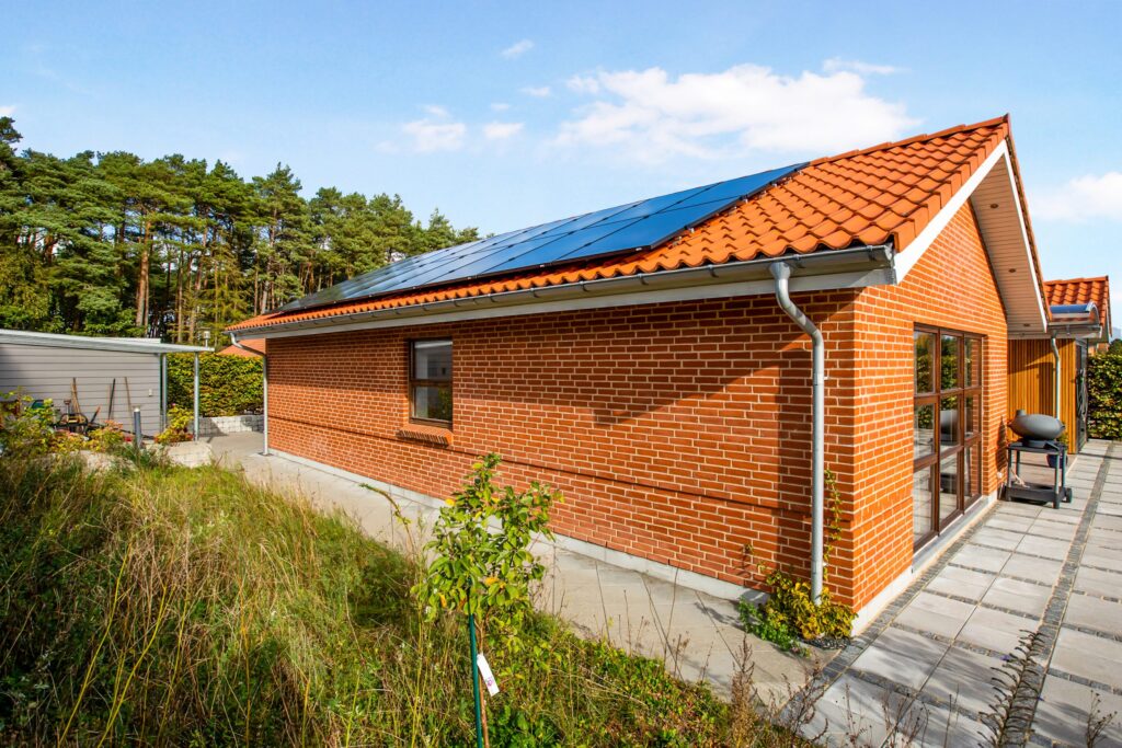 Stort muret hus med rødt tegltag og monterede solpaneler under blå himmel og omgivet af natur.