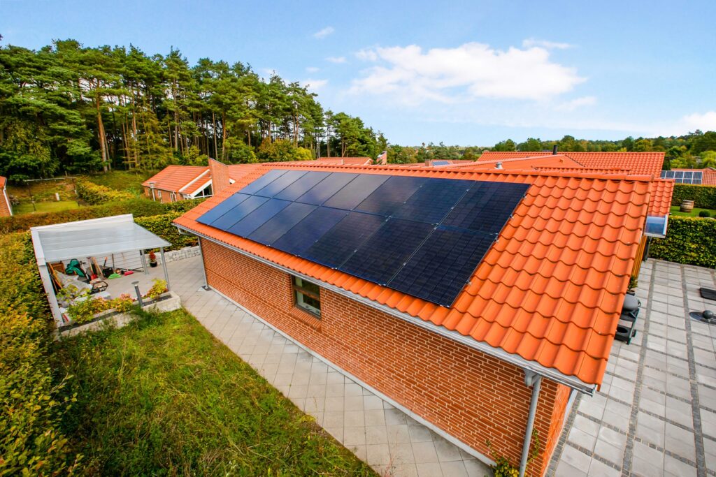 Et lyst, orange tegltag med sorte solcellepaneler på en murstensvilla med grøn beplantning og træer i baggrunden.